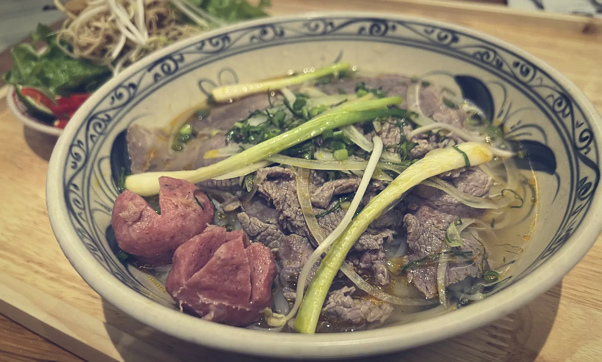 Bowl of Bún bò Huế with garnishes in the background.