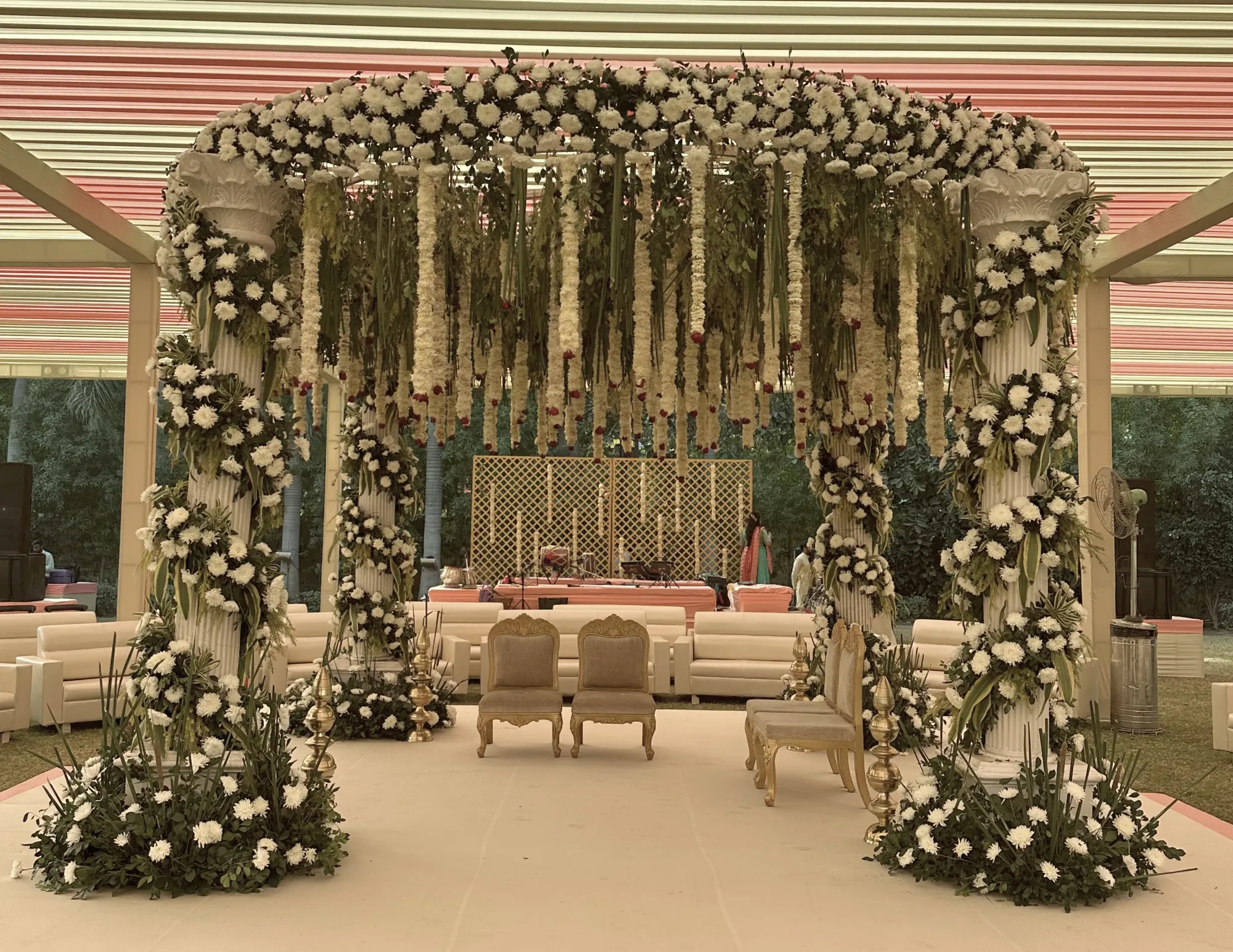White mandap with four columns covered in a garland of white flowers with flowers hanging from the top under a pink and white canopy