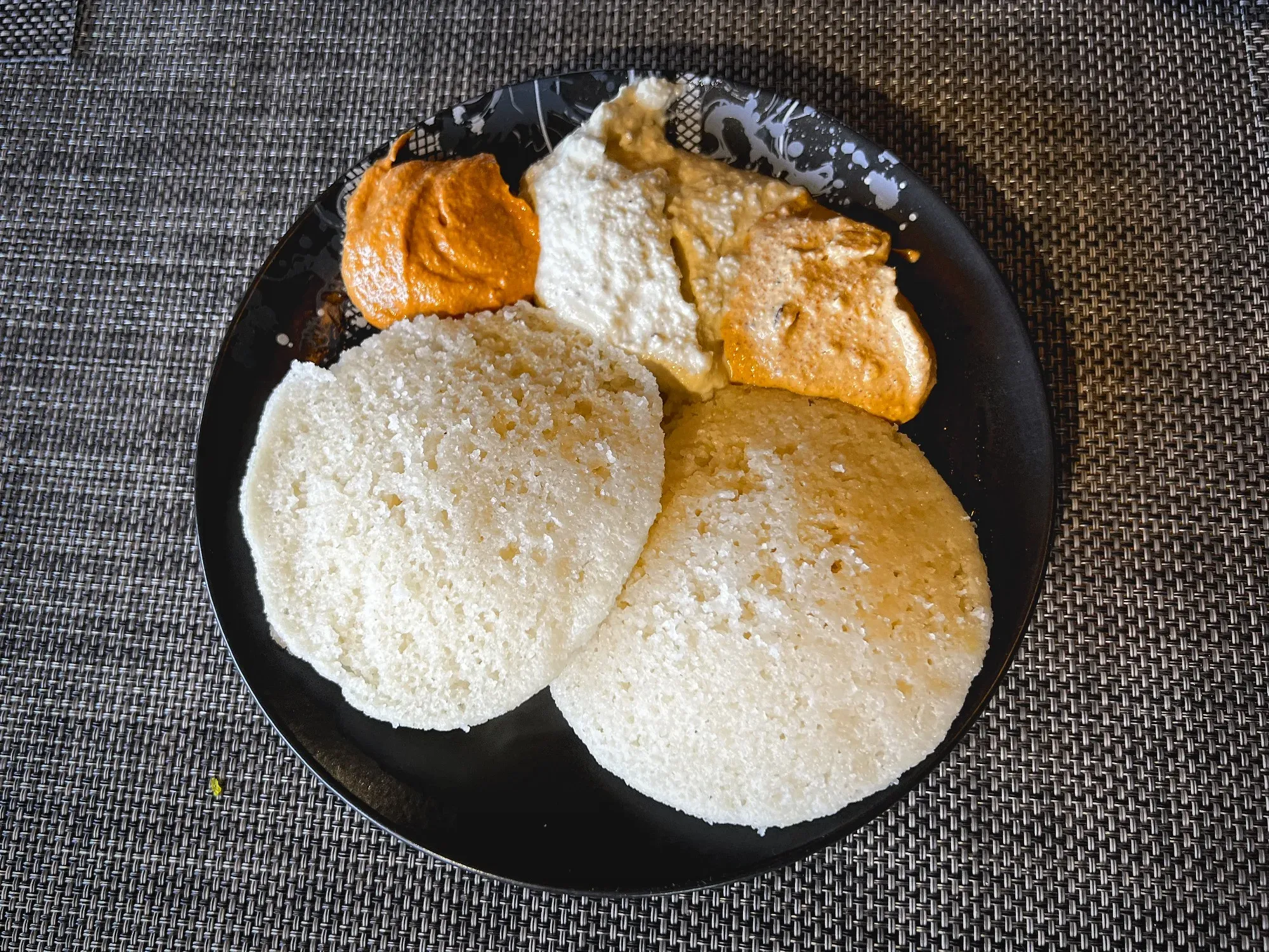 Angled shot of two Idli with three scoops of different side dips on a single plate.