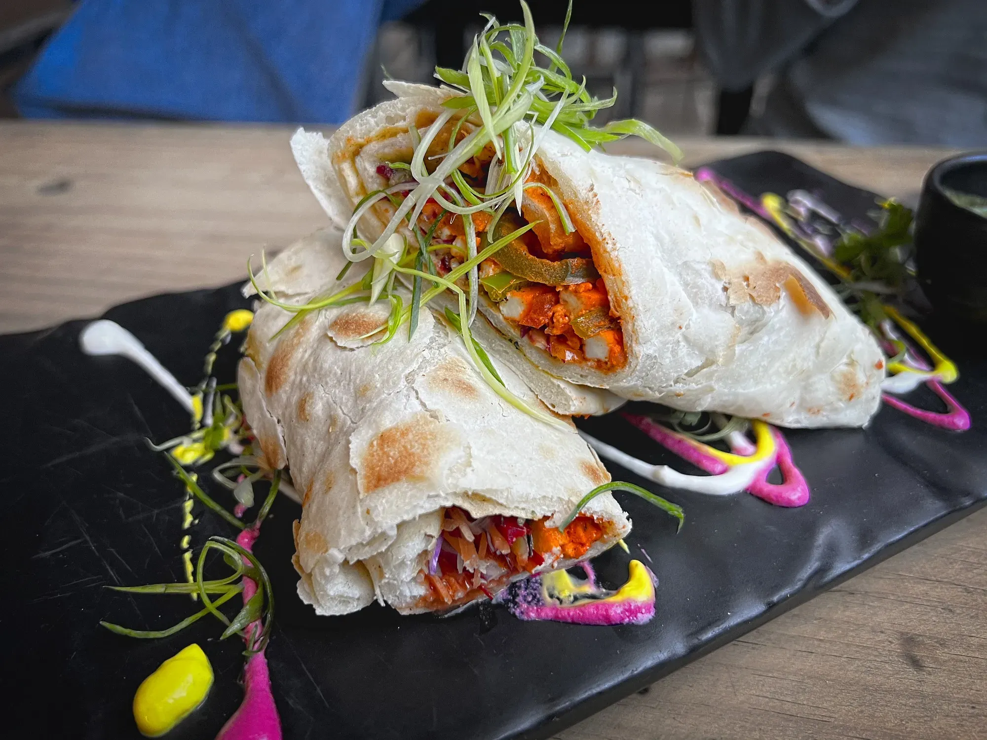 Table shot of a Kathi roll cut in half with pink, yellow, and white sauce plated in an artistic fashion.