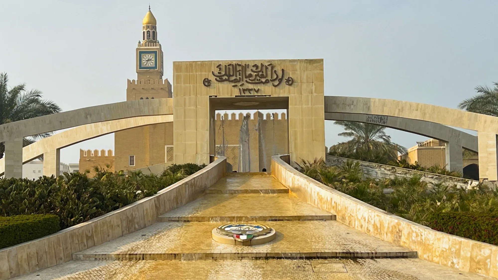 Tan stone fountain with the national emblem in the middle and vegetation on the sides