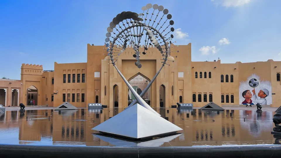 Round fountain with silver geometric figure in the center placed in front of tan buildings with a mural